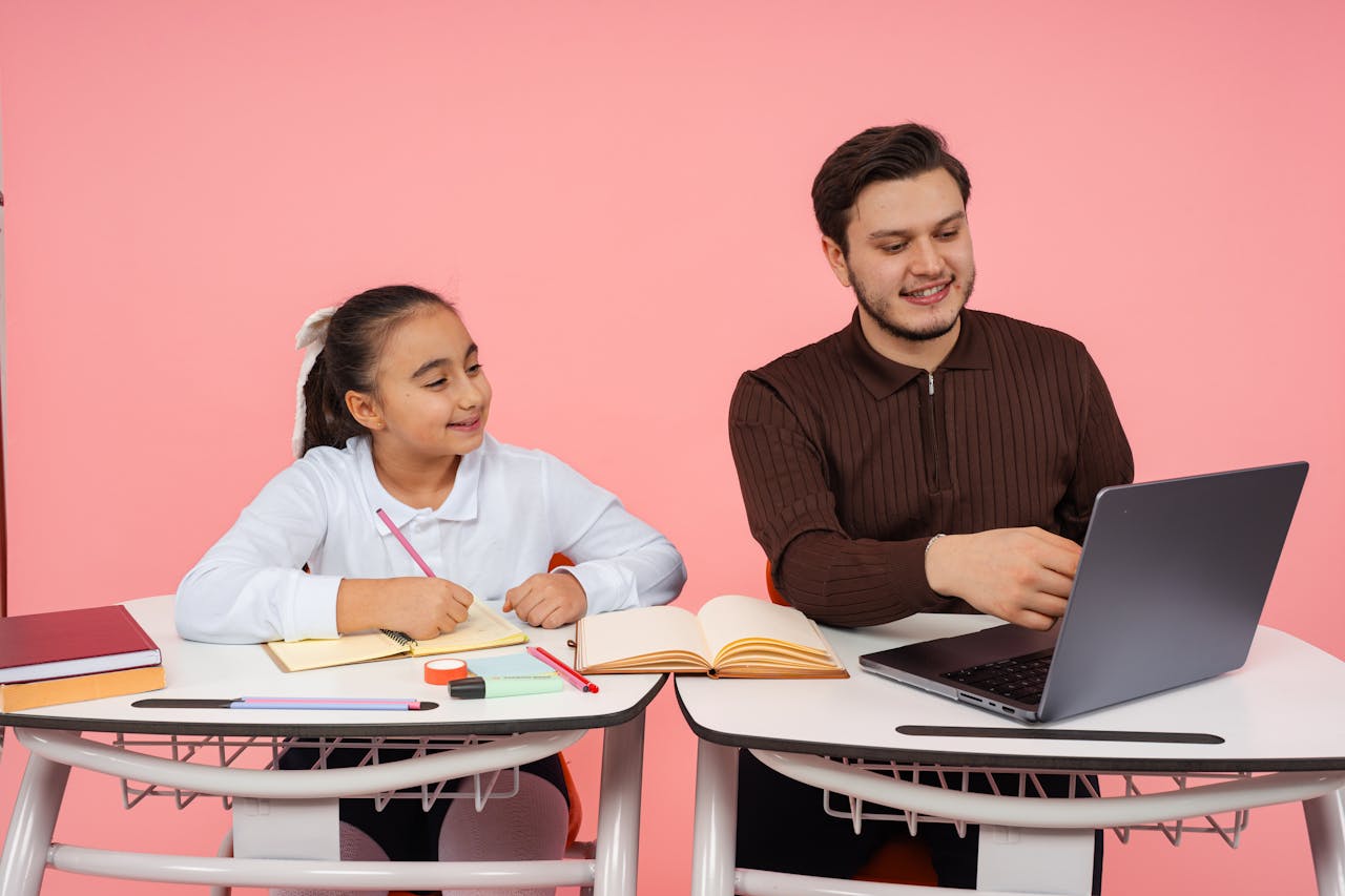 A teacher guides a student in a classroom with books and a laptop on a pink background.